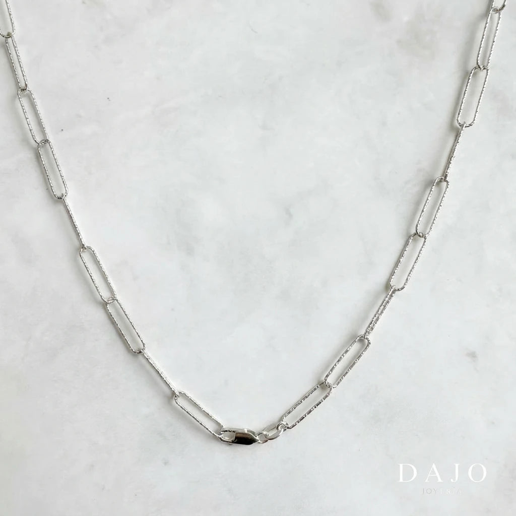 Esta cadena de plata .925 está compuesta por eslabones rectangulares alargados de 5 mm de ancho, formados con finas barras facetadas que tienen un acabado diamantado; un tipo de pulido con textura que refleja la luz en múltiples direcciones.
A pesar de su volumen visual, es una cadena ligera y cómoda para uso diario. Su diseño transmite elegancia con un giro moderno, ideal para quienes buscan una pieza que combine brillo discreto, sofisticación y originalidad