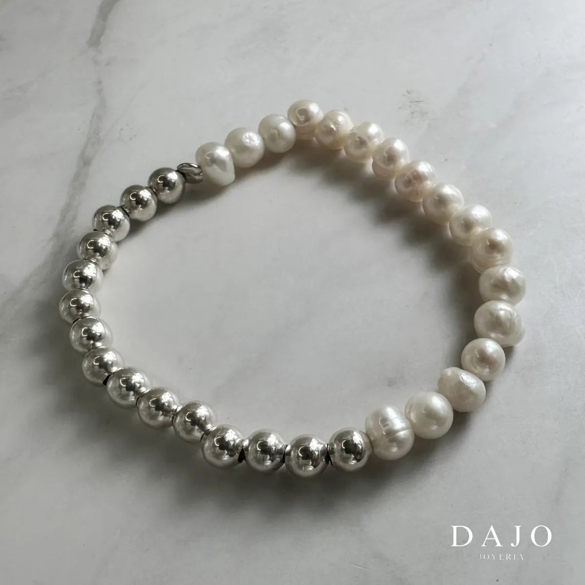 Esta pulsera combina esferas de plata y perlas barrocas, unidas mediante una banda elástica que garantiza comodidad y adaptabilidad.
Las perlas barrocas, conocidas por sus formas únicas e irregulares, aportan un carácter distintivo a la joya, mientras que las esferas de plata añaden un toque de brillo y sofisticación.